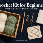 Crochet kit supplies with cream and sage yarn, a wooden hook, scissors, stitch markers and a practice square on a dark background.