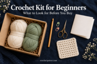 Crochet kit supplies with cream and sage yarn, a wooden hook, scissors, stitch markers and a practice square on a dark background.
